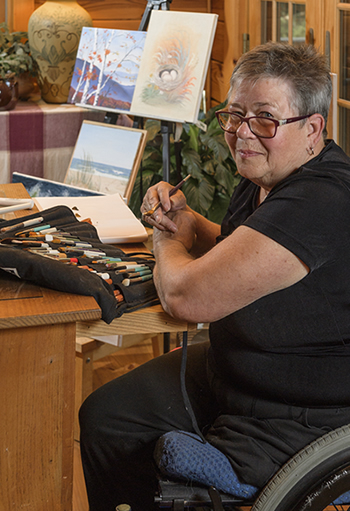 Liisa Trala, Osher Lifelong Learning Institute instructor sitting with brushes in front of a few paintings.