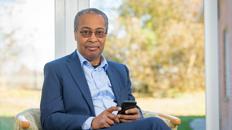 Tony Powell looking forward with smartphone in hand and trees behind window in background