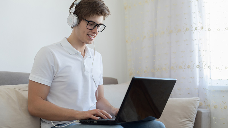 Male student sitting on couch wearing headphones working on laptop