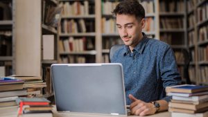 Male high school student on laptop surrounded by books in library