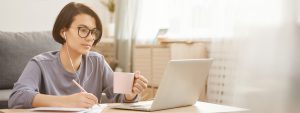 Woman sitting on floor working on laptop with pen in right hand and mug in left hand