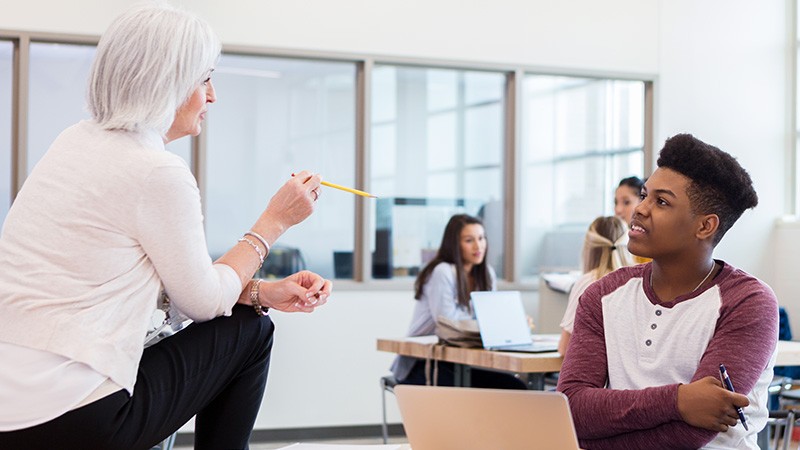 Female teacher sitting on desk talk with male student seated in chair