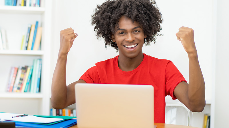 High school student sitting laptop raising two fists in celebration