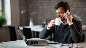 Man talking on phone, drinking coffee, looking at laptop screen