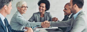 Two businesswomen and three businessmen sitting around a table as two people shake hands to close a deal.