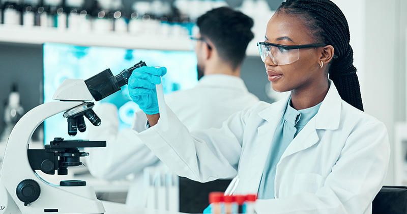 Young woman in a lab coat looking at a test tube, standing in front of a microscope