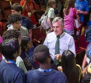 A large group of young students gathers around the governor.