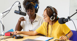 Two teenage girls sitting at desk with two microphones, a laptop and cell phone, recording a podcast