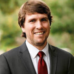John Tadler in a suit on a blurred outdoor background.
