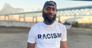 Kevin Martin stands outdoors near a bridge at sunset, wearing a white T-shirt with the word "RACISM" with a slash through it displayed across the front. Martin is a University of Delaware human services student whose educational journey was shaped by support from the ACCESS Center.