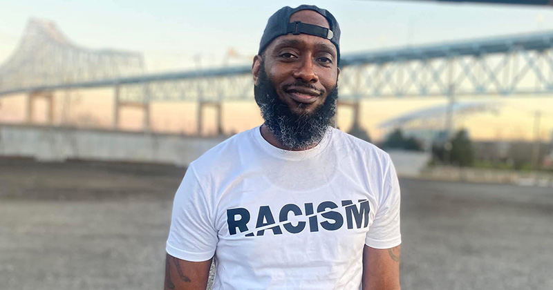 Kevin Martin stands outdoors near a bridge at sunset, wearing a white T-shirt with the word "RACISM" with a slash through it displayed across the front. Martin is a University of Delaware human services student whose educational journey was shaped by support from the ACCESS Center.