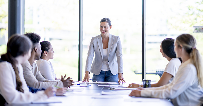 Person standing at the head of a conference table leading a meeting with three colleagues seated on each side.