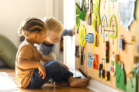 Two young children sit on a hardwood floor and interact with a wall-mounted activity board featuring colorful shapes, gears, laces, and tactile learning elements. Natural light illuminates the scene as the children explore the hands-on educational board together.