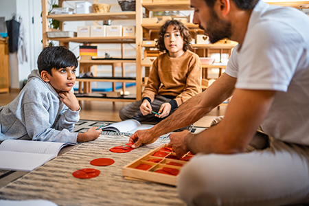 An educator works with learners on a hands-on mathematics lesson using Montessori materials and manipulatives in a classroom setting.