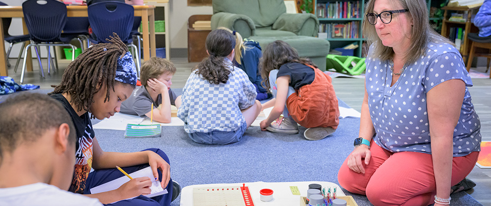 Montessori teacher works with elementary-age students during a classroom learning activity, as students write, collaborate and use Montessori materials on the classroom floor.