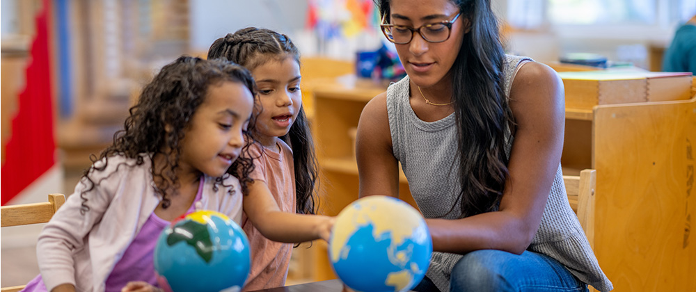 Early childhood teacher working with two young children on a geography lesson that involves globes.