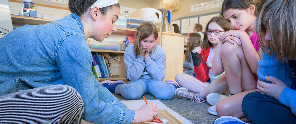 Montessori educator leads a small-group lesson on the classroom floor while elementary-age students observe and participate in a hands-on learning activity.